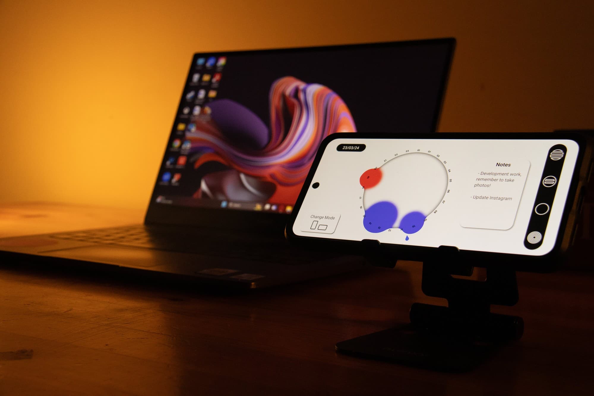 The Memor prototype open on a phone on a stand, showing the fluid clock-calendar with red and blue event circles, beside a laptop on a desk.