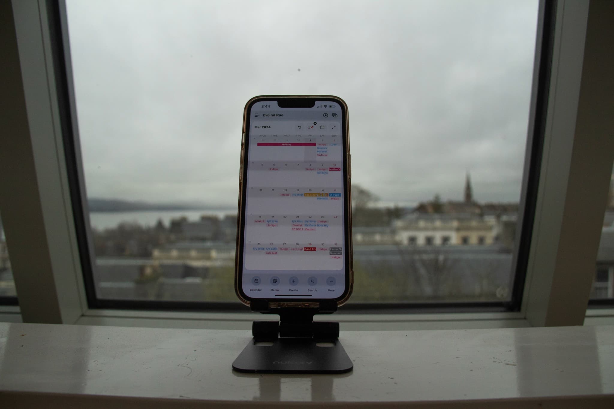 An iPhone on a stand displaying a calendar app, propped on a windowsill overlooking a city view.